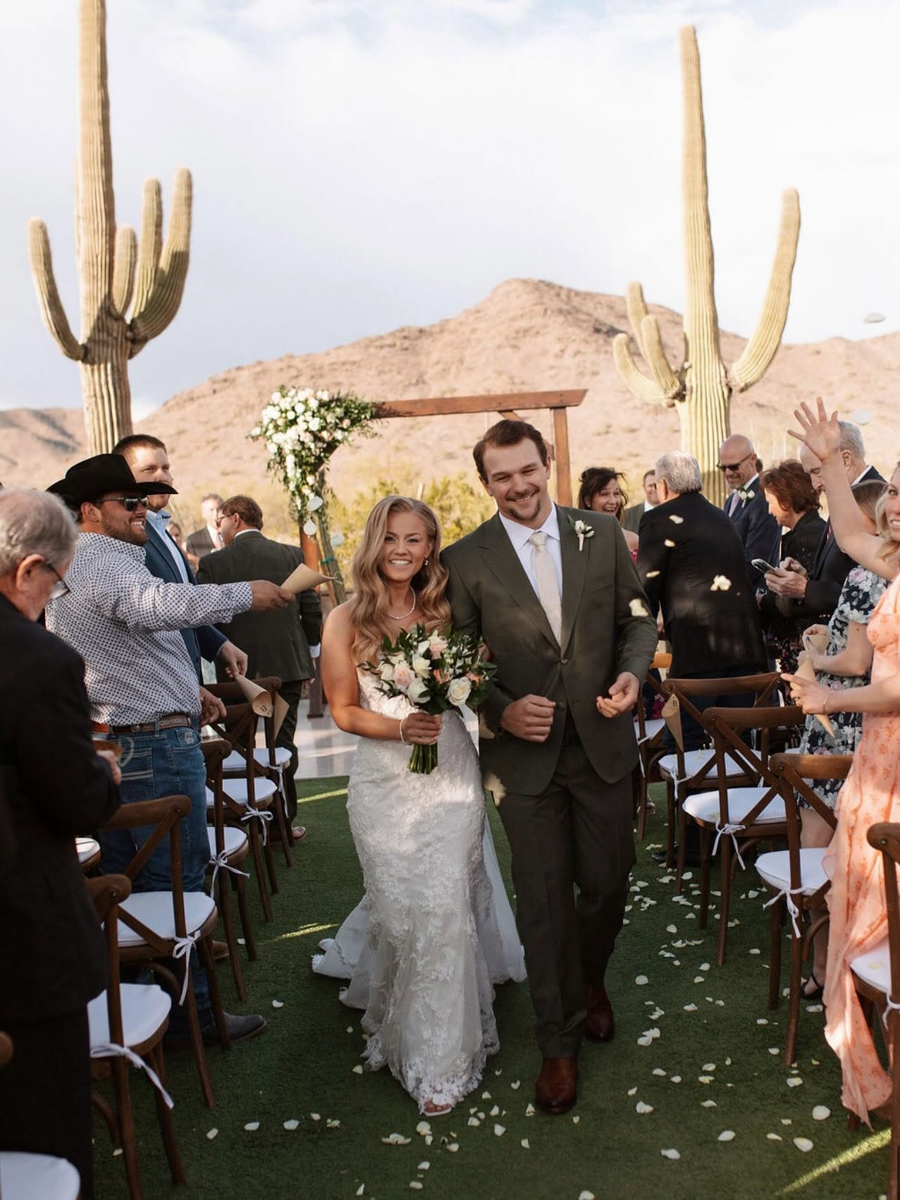 Bride and groom recess down a grass aisle flanked by wooden cross-back chairs after their outdoor desert ceremony, guests tossing petals and cheering. Tall saguaro cacti and a desert mountain backdrop with a wooden arbor draped in white floral garland are visible behind them.