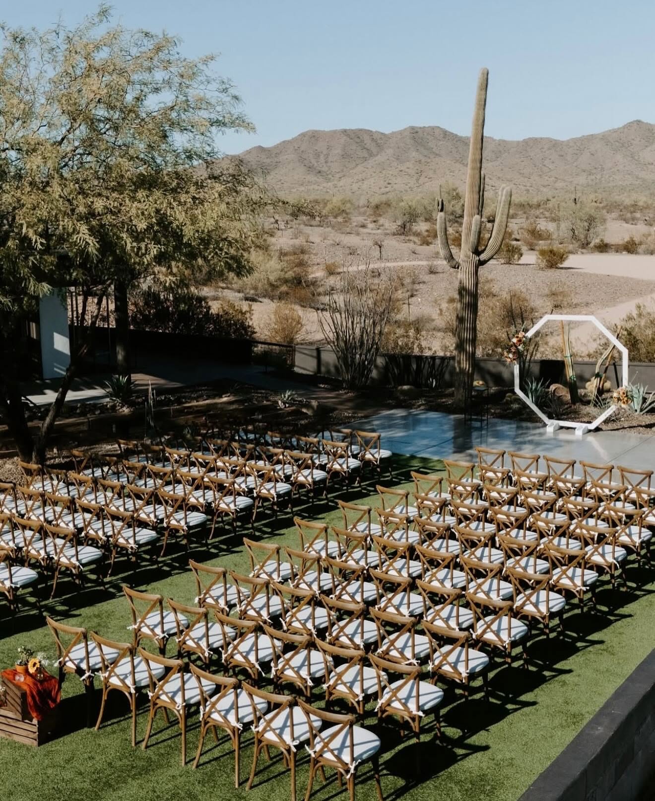 Empty outdoor ceremony setup viewed from above and behind, showing rows of wooden cross-back chairs with white cushions on artificial turf leading to a white geometric (hexagonal) arch decorated with florals. Desert landscaping, a reflective pool/hardscape area, saguaro cactus, and mountain range fill the background under bright daylight.