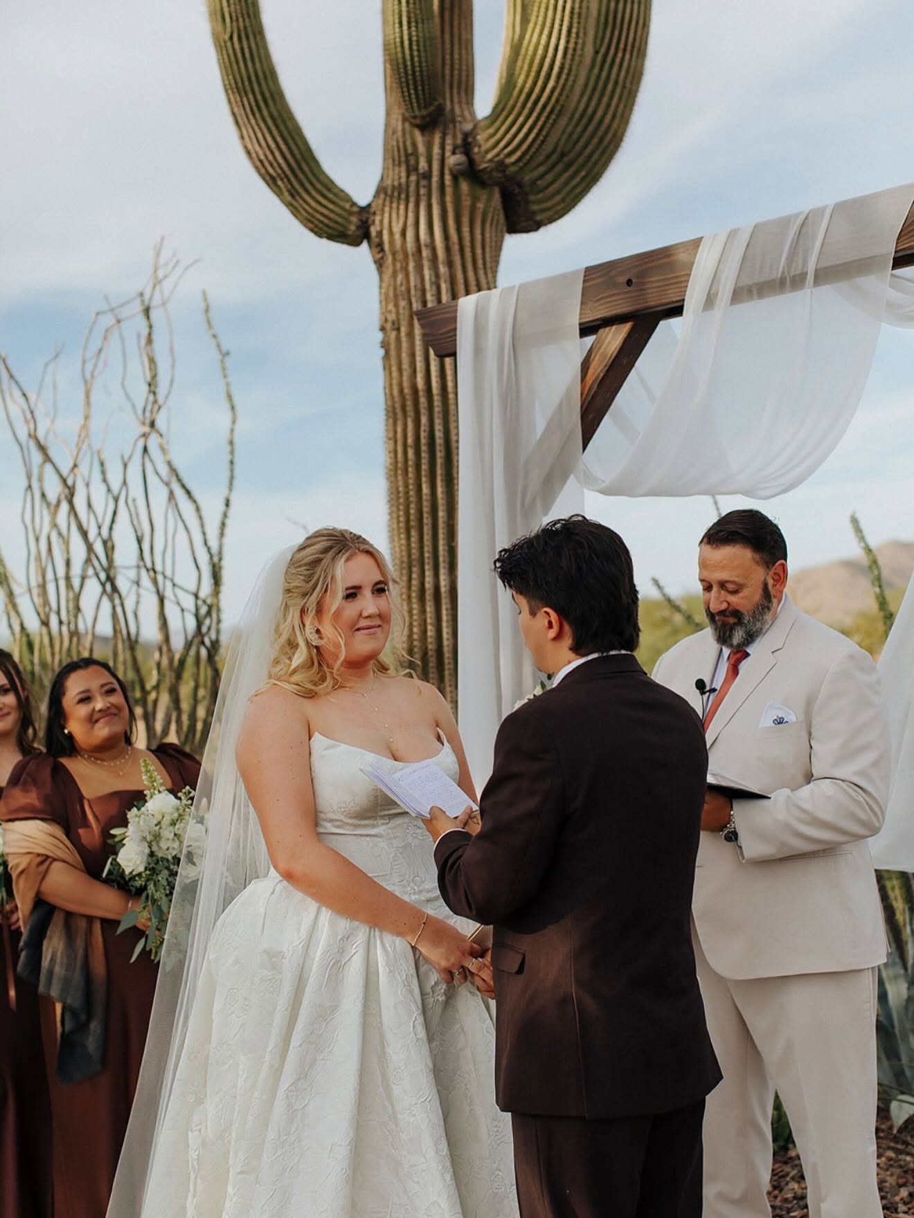Bride and groom hold hands and read vows in front of an officiant under a wooden arbor draped with sheer white fabric, a large saguaro cactus rising directly behind them. Bridesmaids in brown dresses stand to the side; soft daylight suggests late afternoon.