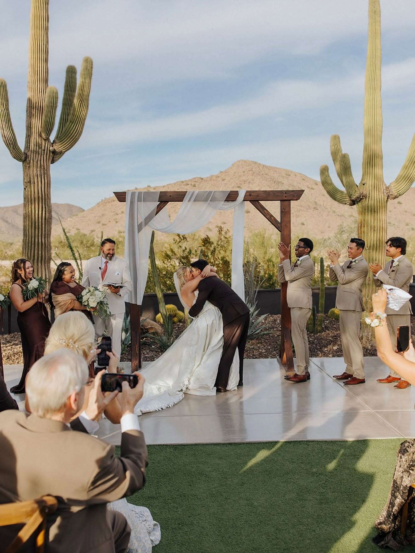 Newlyweds kiss atop a raised concrete platform under a wooden arbor with white drapery, flanked by groomsmen in tan suits applauding and bridesmaids in brown dresses laughing. Saguaro cacti and desert mountains frame the scene with a turf aisle and chairs in the foreground.