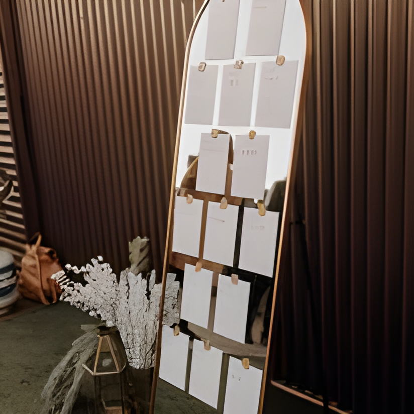 Arched wooden seating chart display propped against a dark corrugated metal wall, with rows of small printed name cards and a dried floral arrangement in a woven vase beside it. Appears to be staged near a reception entrance.