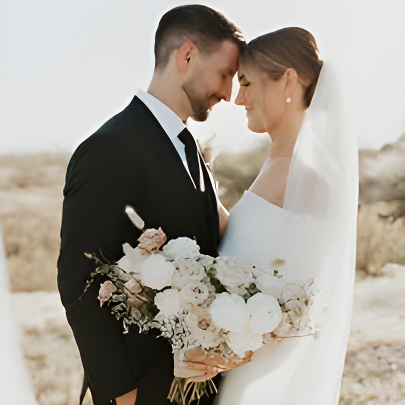 Bride and groom pose forehead-to-forehead outdoors in golden-hour light, with a scrubby desert landscape and distant hills behind them. Bride holds a loose bouquet of white and blush blooms; likely taken somewhere on the venue grounds rather than at a fixed ceremony structure.