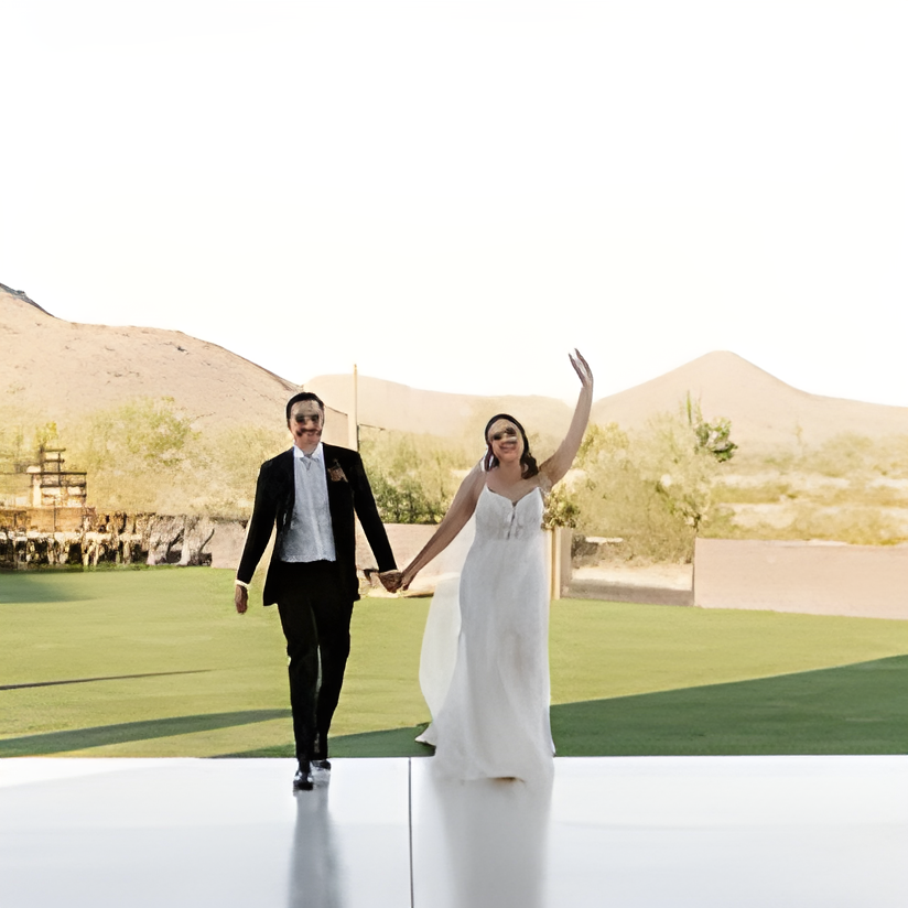 Bride and groom walking hand-in-hand across an open outdoor lawn area with desert mountains in the background, string-light-hung structure visible in the distance. Bright daylight, joyful candid moment.