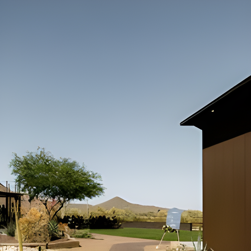 Exterior view of a modern dark-toned building corner with desert landscaping (agave, cacti) in foreground, overlooking a grassy area and mountain in the distance with a small sign and people gathered near a tent.