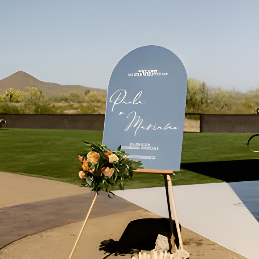 A welcome sign with elegant blue-gray coloring and white script text ('Paola & Maximiliano') stands on a wooden easel beside a paved path, with a small floral bouquet at its base. Manicured green lawn and desert shrubbery are visible under bright daylight.