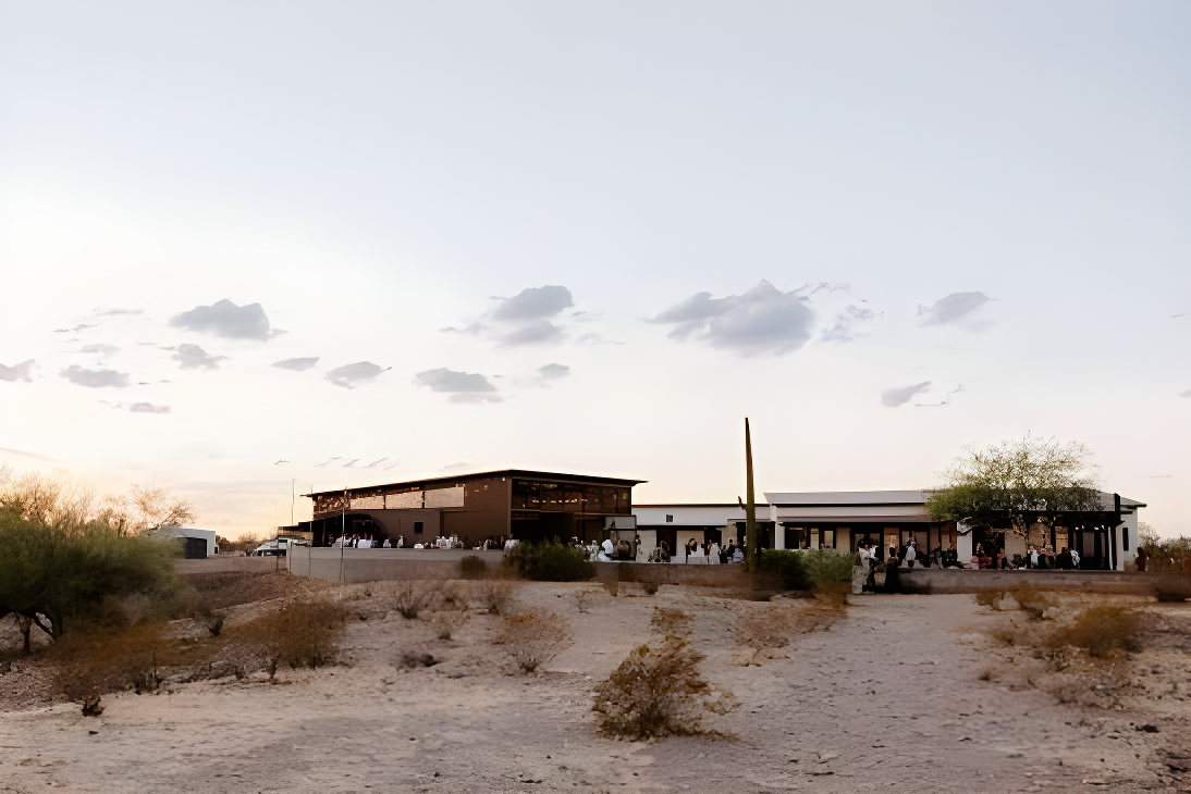Wide exterior view of the venue compound at dusk/golden hour, showing a modern building complex with a dark metal-roofed pavilion structure on the left and a white stucco building with covered patio on the right, guests gathered outside. Sparse desert scrub and a truck in the lot are visible in the foreground.