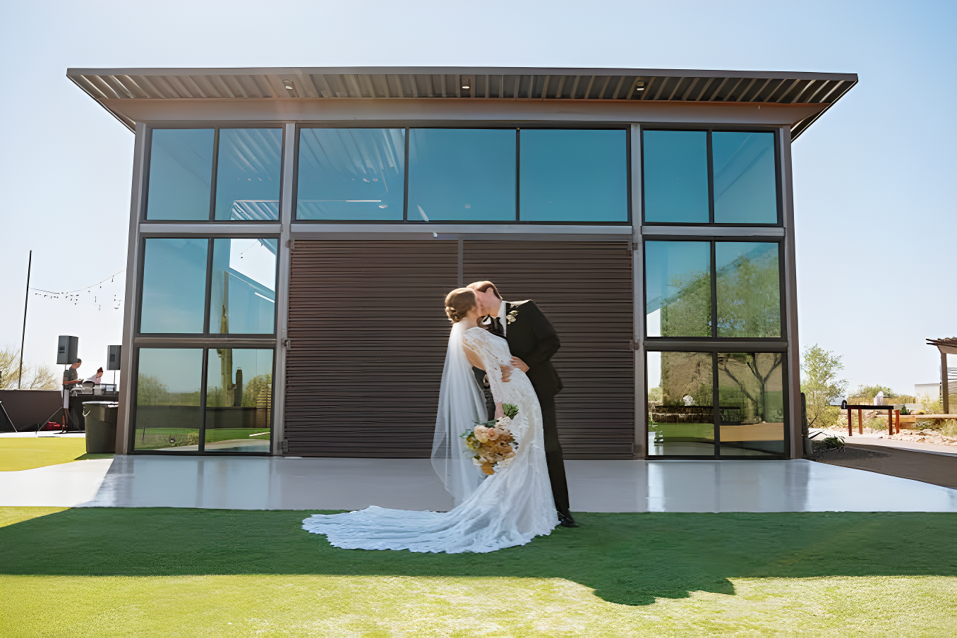 Bride and groom kiss on a lush green lawn in front of a modern industrial-style building with large dark-framed glass windows and a corrugated metal roll-up door, string lights strung along the eave. Clear blue sky and manicured grass suggest a landscaped area adjacent to the reception hall used for portraits.