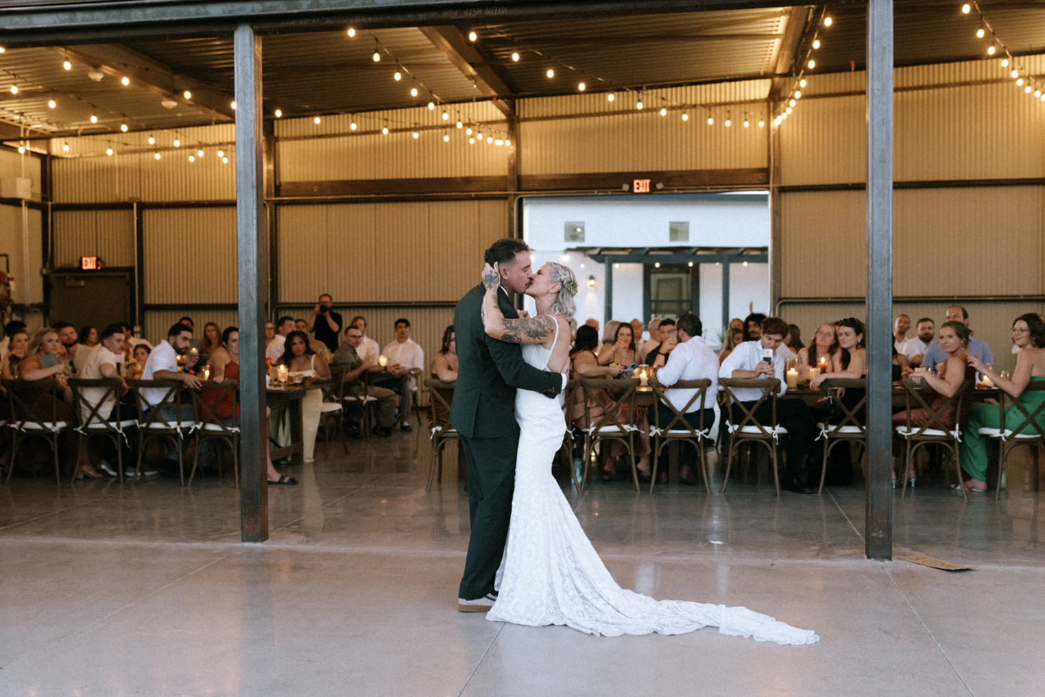 Bride and groom share a first-dance kiss on a polished concrete dance floor inside a large industrial-style barn/hangar reception space with a corrugated metal ceiling and exposed steel beams. Warm string lights crisscross overhead and long farm tables lined with cross-back chairs flank both sides, filled with seated guests. Through an open bay door at the rear, a white stucco building with a covered walkway is visible outside, confirming the indoor reception opens onto the outdoor compound.