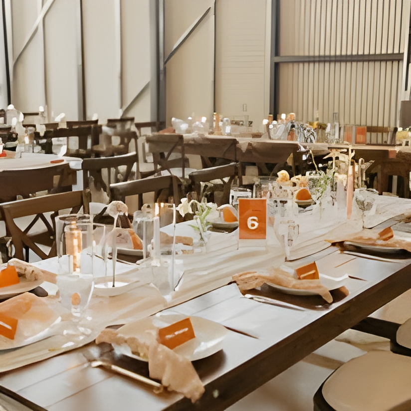 Long wooden farm table set for a reception meal with white linens, orange place cards, glassware, and floral centerpieces, flanked by wooden chairs. Corrugated metal wall visible in background suggests the same industrial barn reception space.