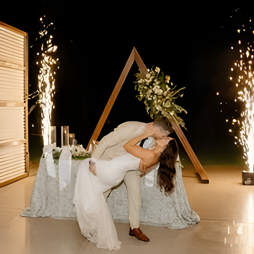 Nighttime reception moment on the venue's outdoor patio: the couple dips into a kiss beside a sequined-linen sweetheart table, framed by a triangular wooden backdrop decorated with greenery and flowers. Cold-spark firework fountains erupt on either side and candles add warm light against the dark night sky.