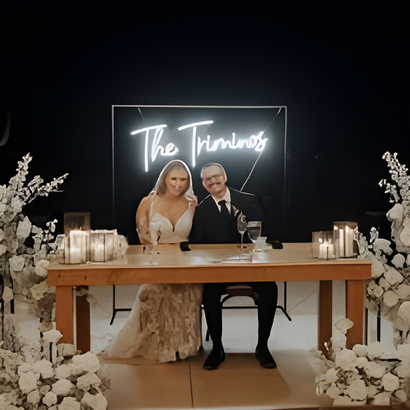 Nighttime sweetheart table shot of a bride and groom seated behind a wooden table flanked by white floral arrangements and pillar candles, with a glowing neon sign reading 'The Trimmins' behind them. Dark surroundings suggest an evening outdoor reception setting.