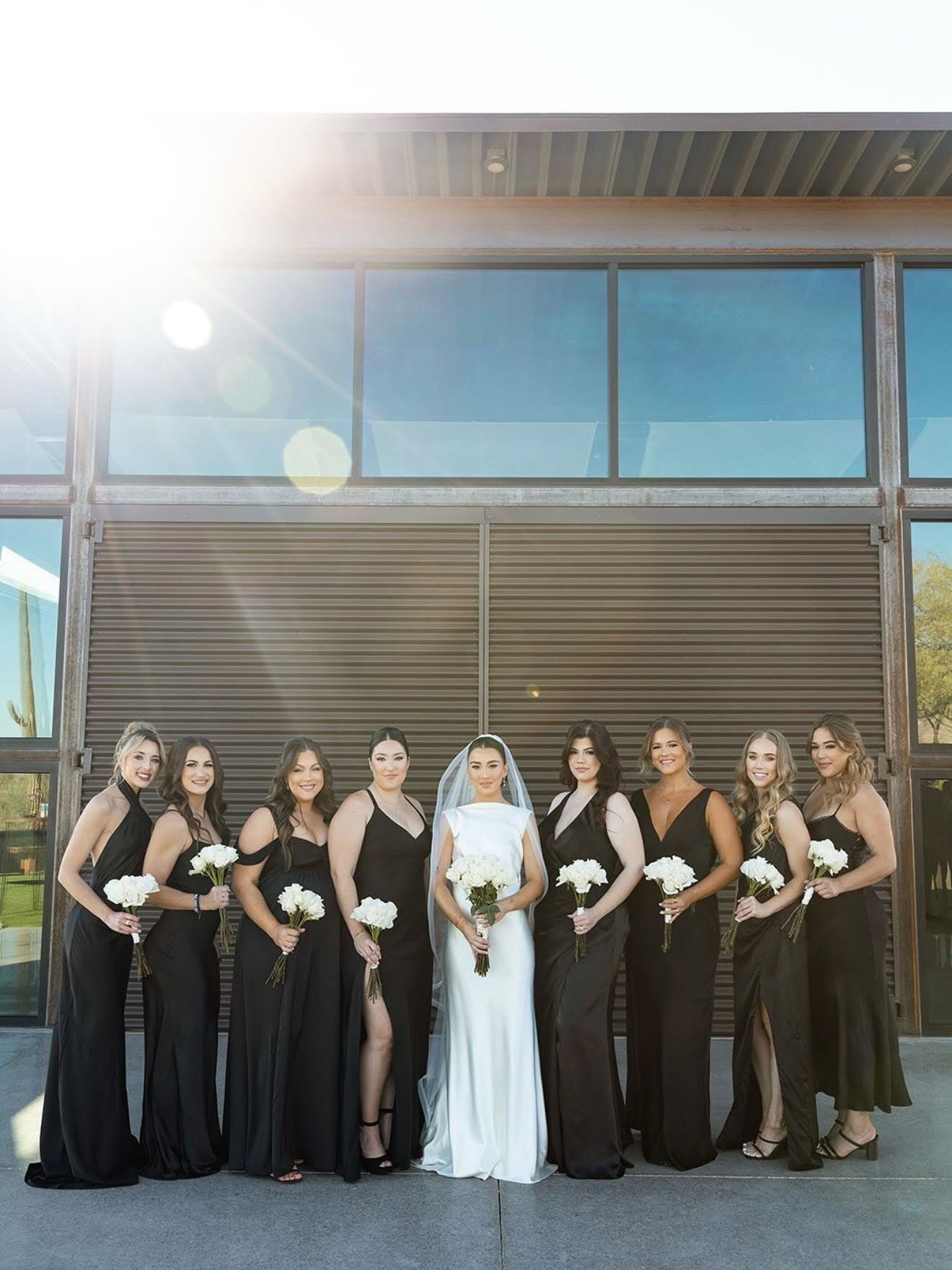 Bride in white gown poses with eight bridesmaids in black dresses, all holding white bouquets, standing on a concrete patio in front of a large industrial-style building with dark corrugated metal panels and big glass windows. Bright sun flare suggests daytime; this appears to be the exterior of the indoor reception building.