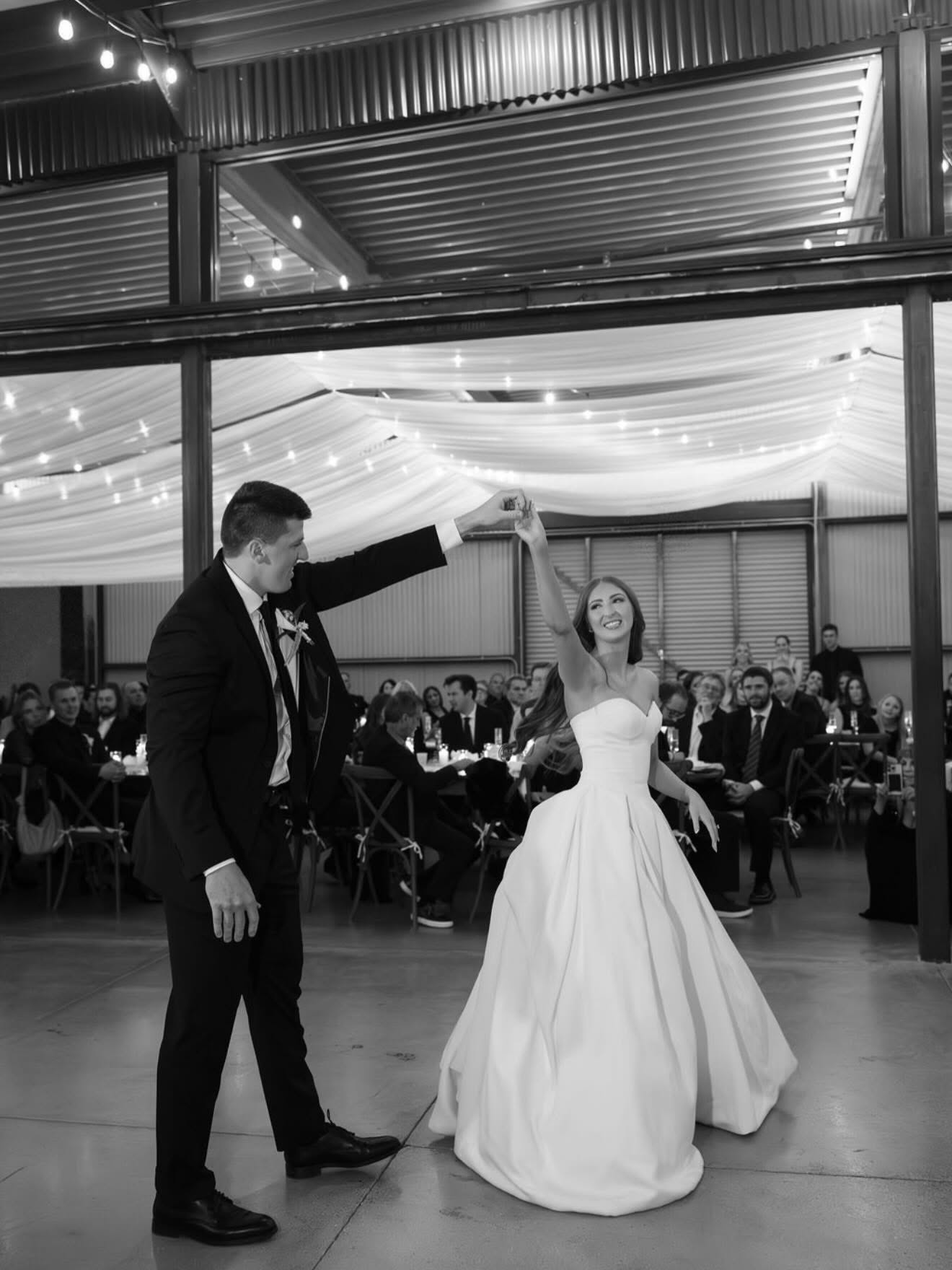 Black-and-white shot of the groom twirling the bride on a concrete dance floor inside a metal-roofed barn/pavilion-style reception building, string lights and draped white fabric overhead, guests seated at long tables in the background. Structural steel beams and corrugated metal walls indicate an enclosed indoor venue.