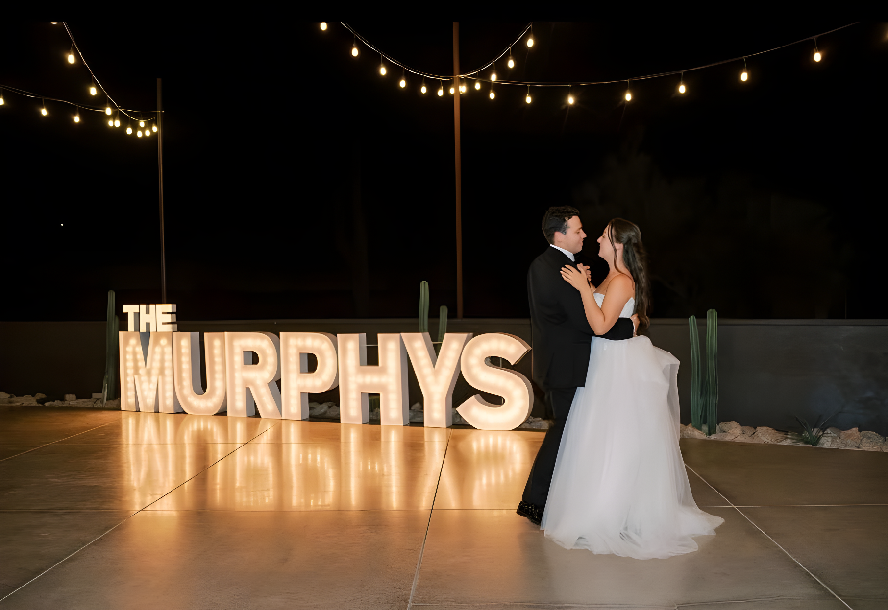 Bride and groom slow dance at night on an outdoor concrete pad next to large marquee-style letters reading 'THE MURPHYS', illuminated by string lights strung overhead. Desert plants and low stone walls edge the space, suggesting an outdoor patio adjacent to the reception building rather than the main indoor hall.