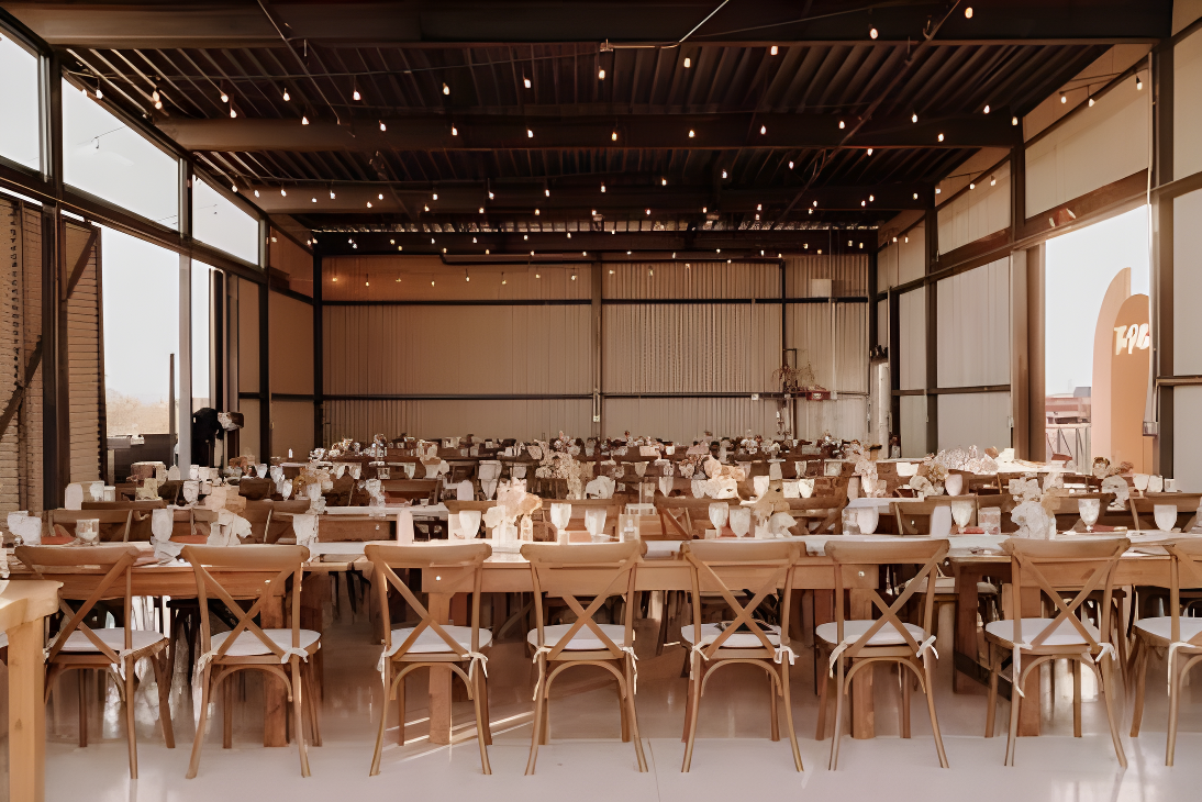 Interior of the reception barn/pavilion set for dinner with long wooden farm tables and cross-back chairs arranged in rows, string lights crisscrossing the exposed steel-beam ceiling. Large glass walls and roll-up doors on the sides let in daylight, and the space appears empty and ready for guests.
