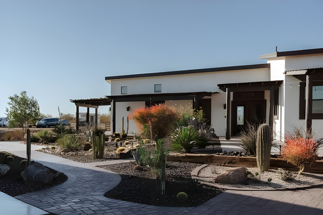 Bride and groom kiss on a concrete pad in front of a large modern reception building with tall glass windows and dark wood accents, string lights along the roofline and desert plants at the base. A wedding DJ setup and speaker are visible to the left, suggesting this pad doubles as the outdoor dance/gathering area beside the indoor hall.